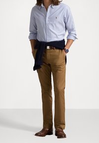 Světle modrá a bílá pruhovaná košile s knoflíky, béžové kalhoty, námořnický svetr uvázaný v pase, hnědé kožené boty, stříbrné hodinky na zápěstí.