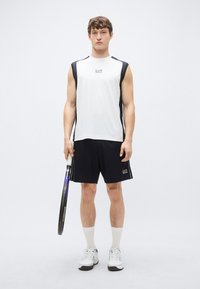 Biele športové tielko bez rukávov s námorníckymi detailmi a logom "EA7", doplnené o čierne šortky. Drží tenisovú raketu a má na nohe biele tenisky.