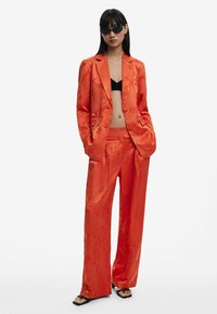 Conjunto de satén naranja con textura floral, que incluye una chaqueta entallada y pantalones de pierna ancha, usado sobre una blusa negra y combinado con sandalias negras.
