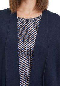 Marinblå cardigan med öppen front, stickad textur, bärs över en mönstrad topp med geometriska former i brunt, blått och krämfärgat.