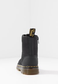 Black ankle boot with textured fabric and smooth heel, yellow stitching, translucent sole, and black-yellow branded pull tab at back.