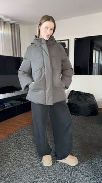 Μπουφάν puffer σε σκούρο γκρι, με κουκούλα, εμπρόσθιες τσέπες και τετράγωνο σχήμα. Συνδυασμένο με μαύρο παντελόνι φαρδιάς γραμμής και μπεζ παπούτσια.