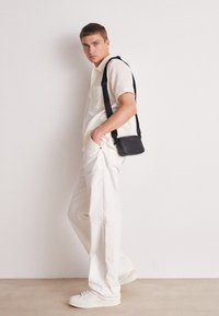 Bolso cruzado negro con textura suave, llevado sobre una camiseta corta de manga corta color crema y jeans blancos holgados, combinado con zapatillas ligeras.