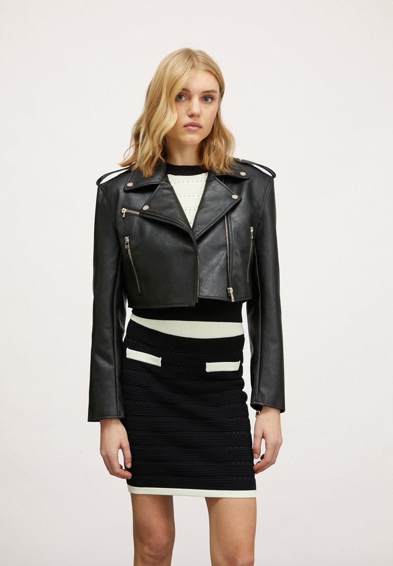 Giacca da moto in pelle nera cropped con hardware argentato, zip dettagliate e epaulettes sulle spalle, abbinata a un vestito aderente bianco e nero.