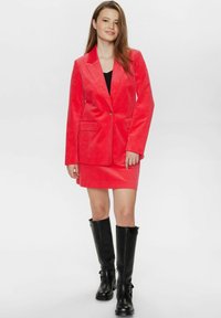 Raudona kordūrojo švarkas su derančia mini sijonu, turi įpjovą ant apykaklės ir priekinėmis kišenėmis, derinama su juodais iki kelių batais.