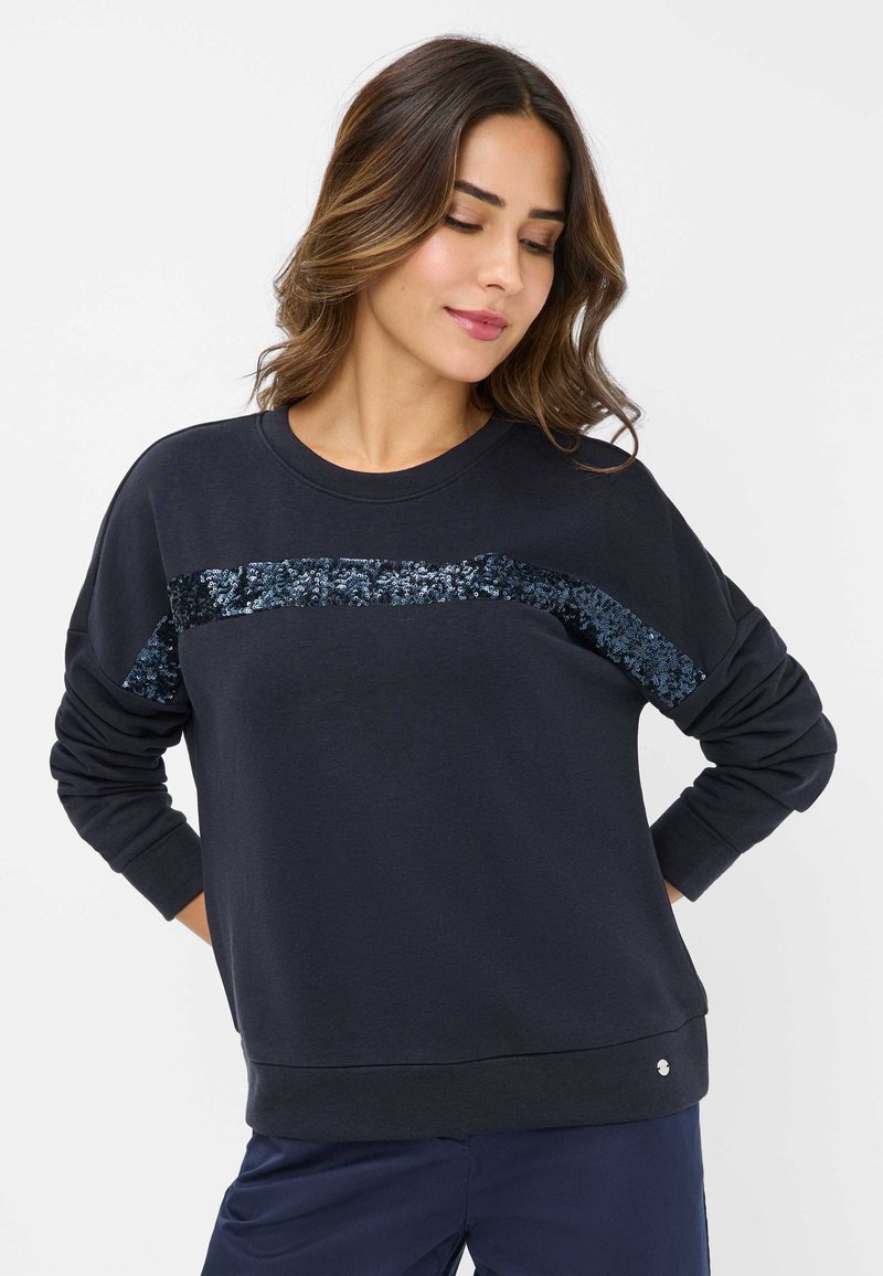 Le sweatshirt bleu marine présente une bande de sequins texturée sur la poitrine, avec des poignets et un ourlet côtelés, fabriqué en un mélange de coton doux.