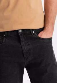 Vaqueros de denim negros con un corte ajustado, que cuentan con una bragueta de botones, bolsillos frontales y un desgaste sutil. Complementados con una blusa color tostado.