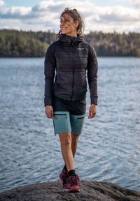 Giacca nera isolante, pantaloni corti neri e verde acqua con tasche con zip, e scarpe da trekking scure. In piedi vicino all'acqua con uno sfondo di bosco.