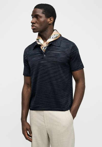 Μαύρος άντρας που φοράει μπλε ναυτικό κοντομάνικο πόλο με ρίγες, μπεζ παντελόνι και μεταξωτό μαντήλι με σχέδια γύρω από το λαιμό του, στέκεται με το ένα χέρι στην τσέπη.