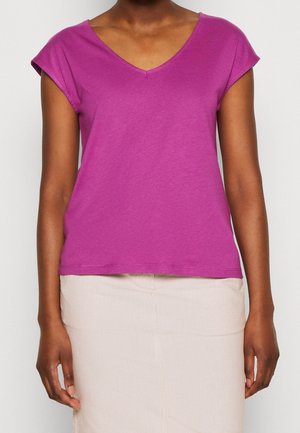 Vrouw draagt een shirt met korte mouwen in magenta met een V-hals en een lichtbeige rok, staand tegen een effen achtergrond.