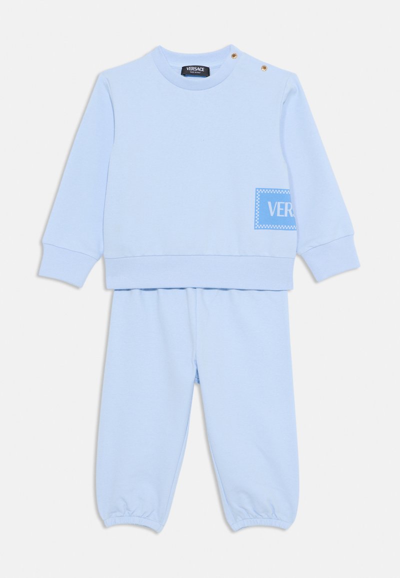 Gaiši zils sporta apģērbs, kas sastāv no kreklā ar noņemamiem pogām un atbilstošām jogger biksēm, abi izgatavoti no mīksta materiāla, ar logo detalizāciju.