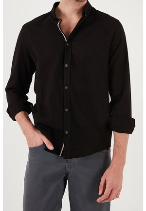 Homme portant une chemise noire à manches longues boutonnée avec les poignets retroussés et un pantalon gris, une main dans la poche, sur un fond uni.