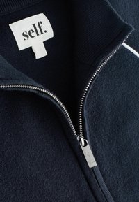 Veste en polaire zippée bleu marine avec une fermeture éclair argentée dotée d'une étiquette distincte portant l'inscription "self." et des détails de col côtelé sur une texture de tissu doux.
