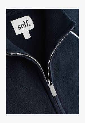 Jachetă fleece bleumarin cu fermoar argintiu și etichetă distinctă cu inscripția "self." și detalii cu guler reiat pe o textură moale a materialului.