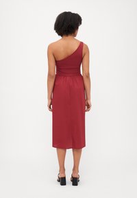Vestido asimétrico de un solo hombro en un rojo intenso, con un cuerpo ajustado y cintura fruncida, y falda fluida hasta la rodilla. Tela suave, diseño sencillo.