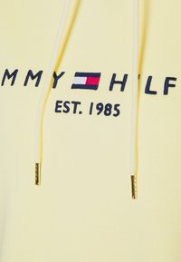 Sudadera de color amarillo claro con texto bordado en marino "TOMMY HILFIGER EST. 1985" y cordones con puntas doradas. Textura de tejido suave.