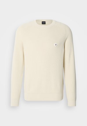 Kremowy sweter z prążkowanej tkaniny, z okrągłym dekoltem, długimi rękawami oraz małym okrągłym logo na lewej piersi.