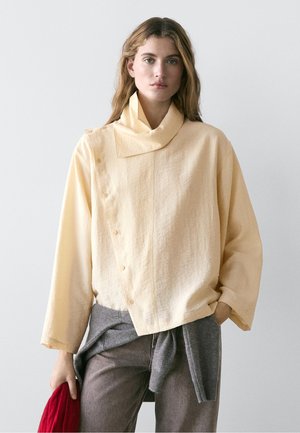 Frau trägt ein lockeres, hellgelbes Langarmshirt mit Knopfdetails und einen grauen Pullover, der um graue Hosen gebunden ist, und hält einen roten Gegenstand.