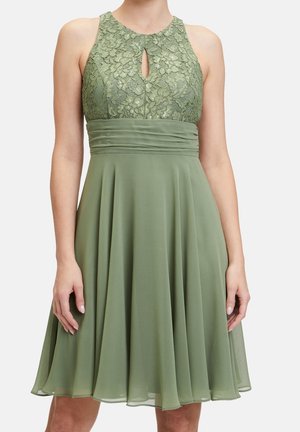 Robe sans manches vert olive avec corsage en dentelle, encolure en trou de serrure, large ceinture froncée et jupe fluide en chiffon longueur genoux portée par une personne debout.