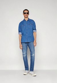 Džinsinis marškiniai vidutinės mėlynos spalvos, su sagomis priekyje, dvi krūtinės kišenės ir suvynioti rankogaliai. Aptempti džinsai su dėvėjimosi detalėmis. Balti sportbačiai.