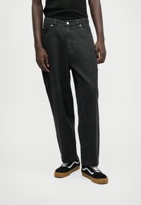 Μαύρα τζιν με χαλαρή γραμμή, σχεδίαση πέντε τσέπες και ασημί μεταλλικά στοιχεία. Συνδυασμένα με μαύρα και άσπρα αθλητικά παπούτσια. Απλό φόντο.