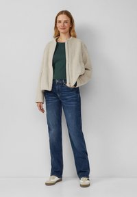 Hellbeige Zip-Up-Jacke über einem dunkelgrünen Shirt, kombiniert mit blauen Jeans und beigen Sneakers mit weißen Akzenten. Lässig, entspannte Passform.
