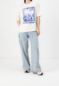 Krämfärgad t-shirt med blå palmträdsgrafik och texten "BILLABONG", ihop med ljusblå vida jeans och gråsvarta sneakers.