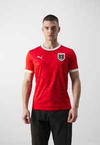 Crvena Puma nogometna majica s teksturiranim uzorkom, bijelim ovratnikom i emblemom na prsima. Ima kratke rukave i prilagođeni dizajn.