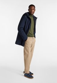 Giacca impermeabile blu con cappuccio, camicia verde a maniche lunghe, pantaloni beige e sneakers blu con accenti azzurri e suole di gomma.