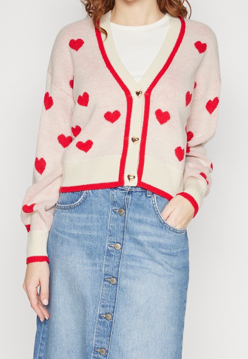 Cardigan crema con motivo a cuori rossi e bordo rosso, con scollo a V, maniche lunghe e bottoni dorati a forma di cuore. Indossato sopra una maglietta bianca.