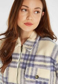 Fleece jack met een crèmekleurige basis, voorzien van een paars en blauw ruitenpatroon, met ritssluiting en een borstzak met knopendetail.