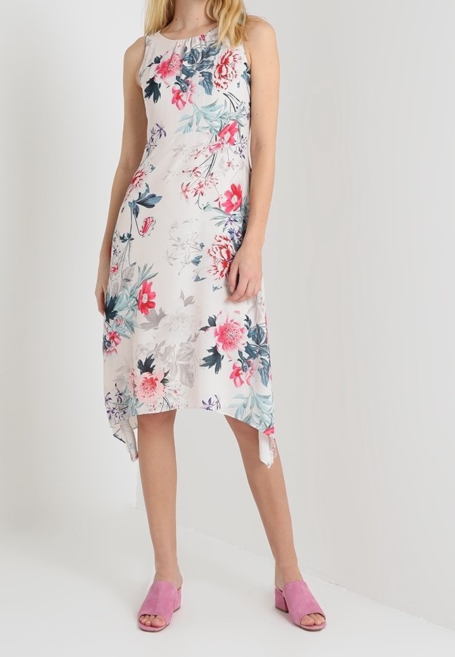 Robe blanche sans manches avec imprimés floraux roses et verts, ourlet asymétrique, portée avec des sandales roses à talons blocs ouverts aux orteils.