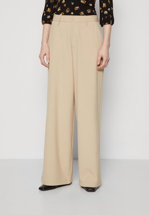 Kobieta w beżowych szerokich spodniach z przodu z guzikami i czarnej bluzce w kwiaty, stojąca na tle jednolitego białego tła.