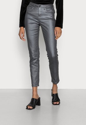 Woman wearing metallic gray slim-fit pants with high waist and black open-toe heeled mules, standing against plain light background.