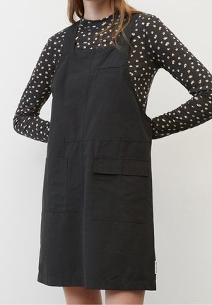 Femme portant une robe tablier noire sans manches avec de grandes poches avant, sur une chemise noire à manches longues avec un petit imprimé floral blanc.
