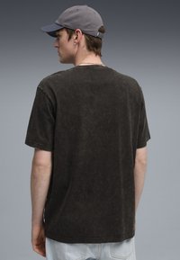 Zwarte katoenen oversized t-shirt met korte mouwen, met een gewassen textuur. Model draagt lichtblauwe jeans en een grijze pet, gezien van achteren.