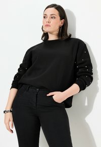 Felpa nera con dettagli in perle sulle maniche, scollatura rotonda e vestibilità comoda, abbinata a jeans neri e un bracciale in argento.