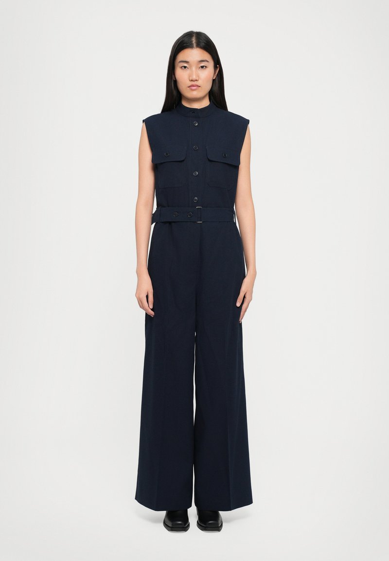 Navy sleeveless jumpsuit with a stand-up collar, button closure, two chest pockets, and a belted waist. Wide-leg design, tailored fit.