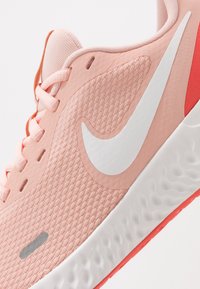 Scarpa da ginnastica rosa chiaro con tomaia testurizzata, logo swoosh bianco e dettagli rossi. Presenta lacci piatti e una suola spessa bianca con motivo a onde.