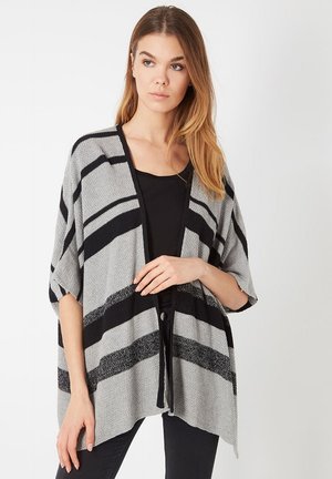 Joven mujer con cabello largo que lleva un cárdigan de rayas gris y negro abierto por delante, sobre una camiseta negra y pantalones negros.