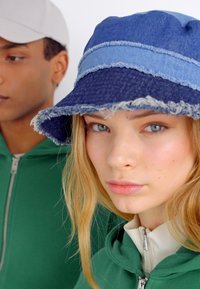 Chapeau seau en denim avec un bord effiloché, présentant un dessus bleu et une base bleu plus foncé ; texture douce. Le modèle porte un sweat à capuche zippé vert.