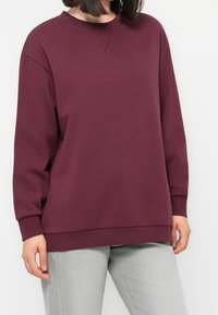 Femme portant un sweat-shirt bordeaux uni à manches longues et un jean gris clair, les mains détendues le long du corps.