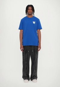 Man stands facing forward wearing a blue T-shirt with white logo, black wide-leg jeans, and white sneakers against a plain white background.