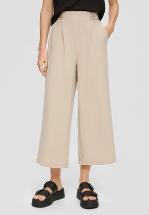 Femme portant un pantalon beige large et court avec des boutons sur le côté et des sandales à plateforme noires, mains dans les poches devant un fond uni.