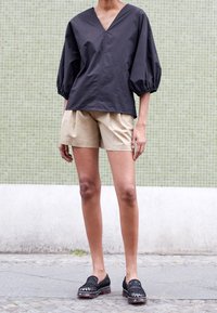 Top nero con scollo a V e maniche a palloncino abbinato a pantaloni corti beige; la modella indossa scarpe nere con dettagli rigati; sfondo a piastrelle verdi.