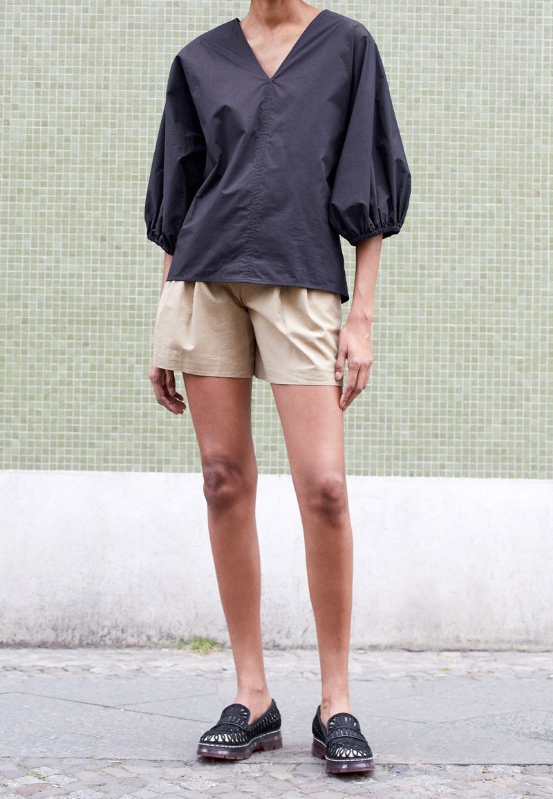 Top nero con scollo a V e maniche a palloncino abbinato a pantaloni corti beige; la modella indossa scarpe nere con dettagli rigati; sfondo a piastrelle verdi.