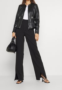 Černá kožená bunda s přiléhavým střihem, kombinovaná s černými širokými kalhotami s vysokým pasem a postranními rozparky. Doplňky zahrnují malou černou kabelku.