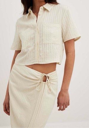 Kvinde iført en cremefarvet kortærmet knap-op crop top med brystlomme og en matchende slå-om nederdel bundet i taljen.