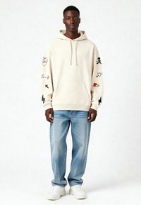 Felpa beige con cappuccio e grafiche sulle maniche, tasca frontale, abbinata a jeans azzurri chiari e sneakers bianche.