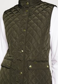 Olivgrön vadderad väst med hög krage, tryckknappar och två framfickor. Slät textur och lätt design.